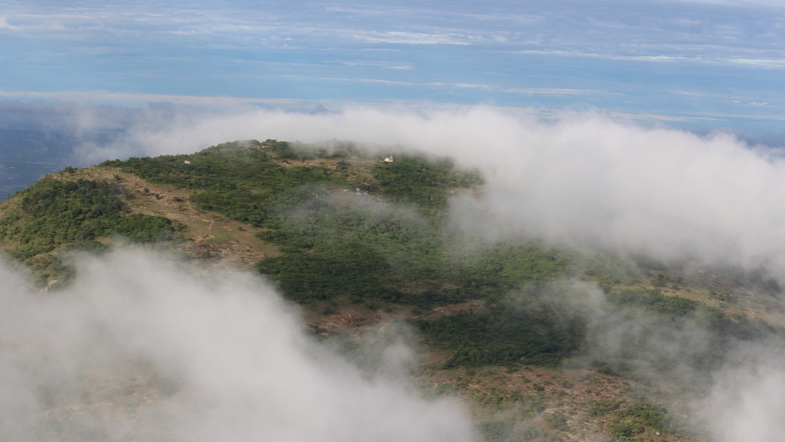 A green, scrub-covered hill rising out of low clouds, seen from the viewpoint at Nandi Hills