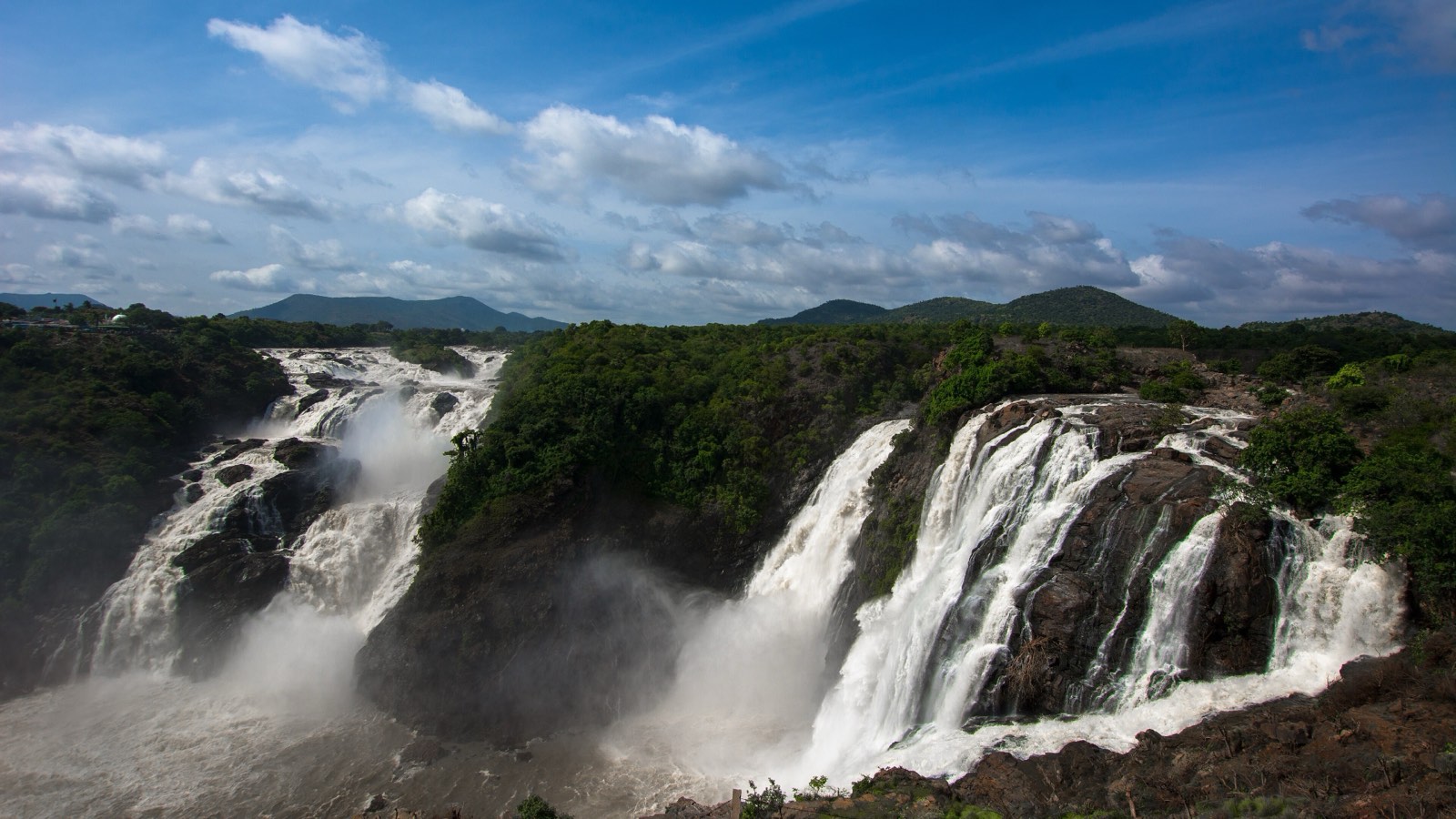 The Kaveri pouring over dark rock in two wide, foaming waterfalls, with forested hills behind