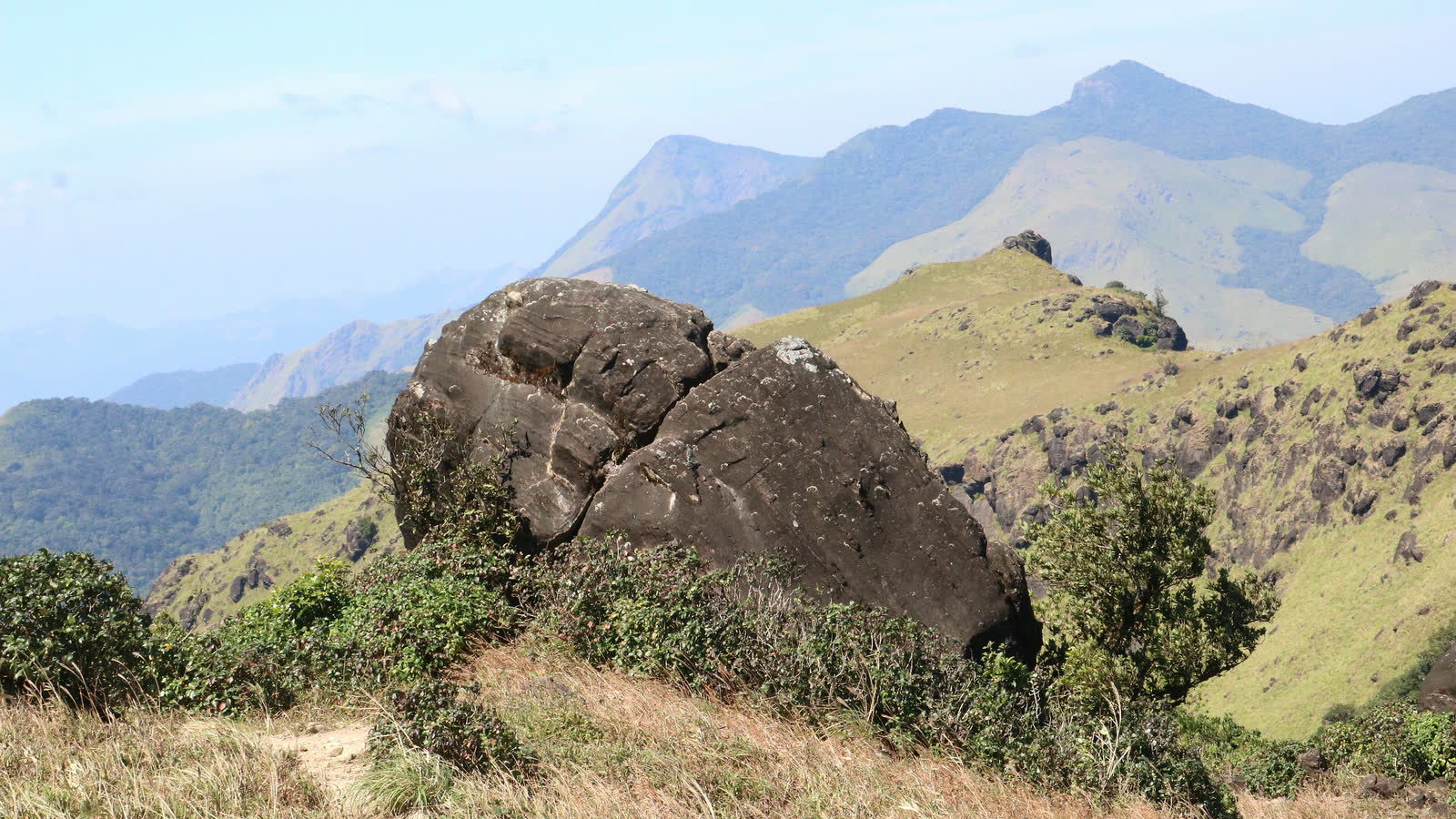 A large rounded boulder on a grassy ridge of Thadiandamol in Coorg, with dry grass in front and layers of blue hills behind