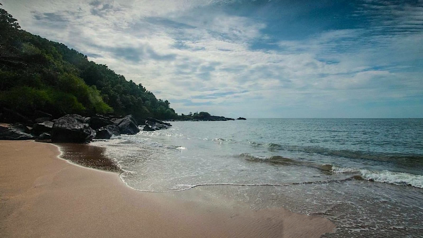 An empty curve of sand at Half Moon Beach in Gokarna, with dark rocks and a forested headland at the far end and small waves coming in
