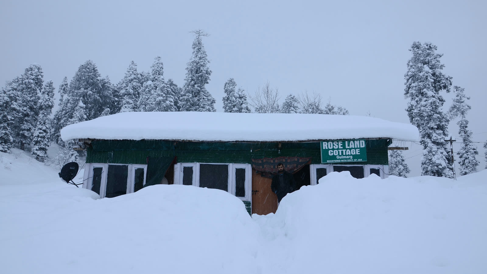 A small cottage in Gulmarg buried to its windows in deep snow, with snow-covered fir trees behind it under a white sky