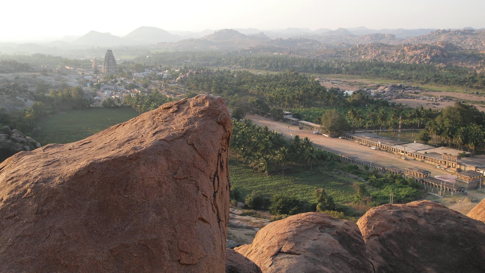 Hampi seen from the top of Matanga Hill, with large orange granite boulders in front and the tall gopuram of the Virupaksha temple among palm groves below