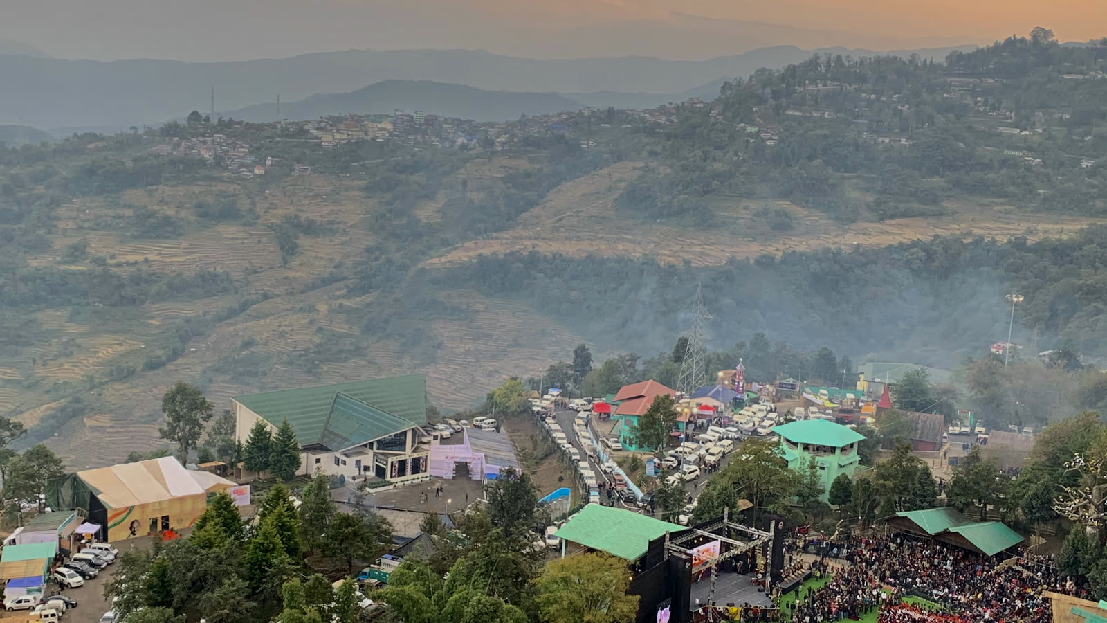 Kisama Heritage Village from above during the Hornbill Festival, with green-roofed buildings, a crowd around the main arena and hazy Naga hills behind