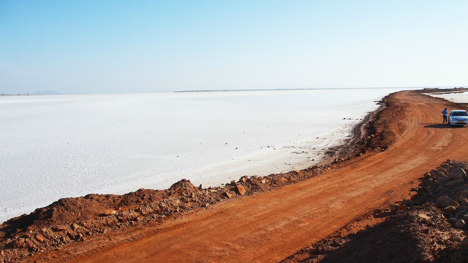 A red dirt road running along the edge of the flat white salt of the Rann of Kutch under a clear sky, with a car parked far down the road