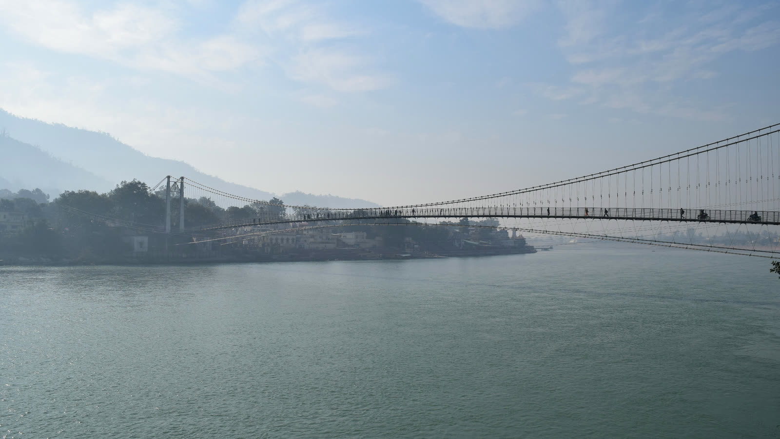 The wide green Ganga at Rishikesh on a hazy morning, with a long suspension bridge crossing the river and forested hills behind