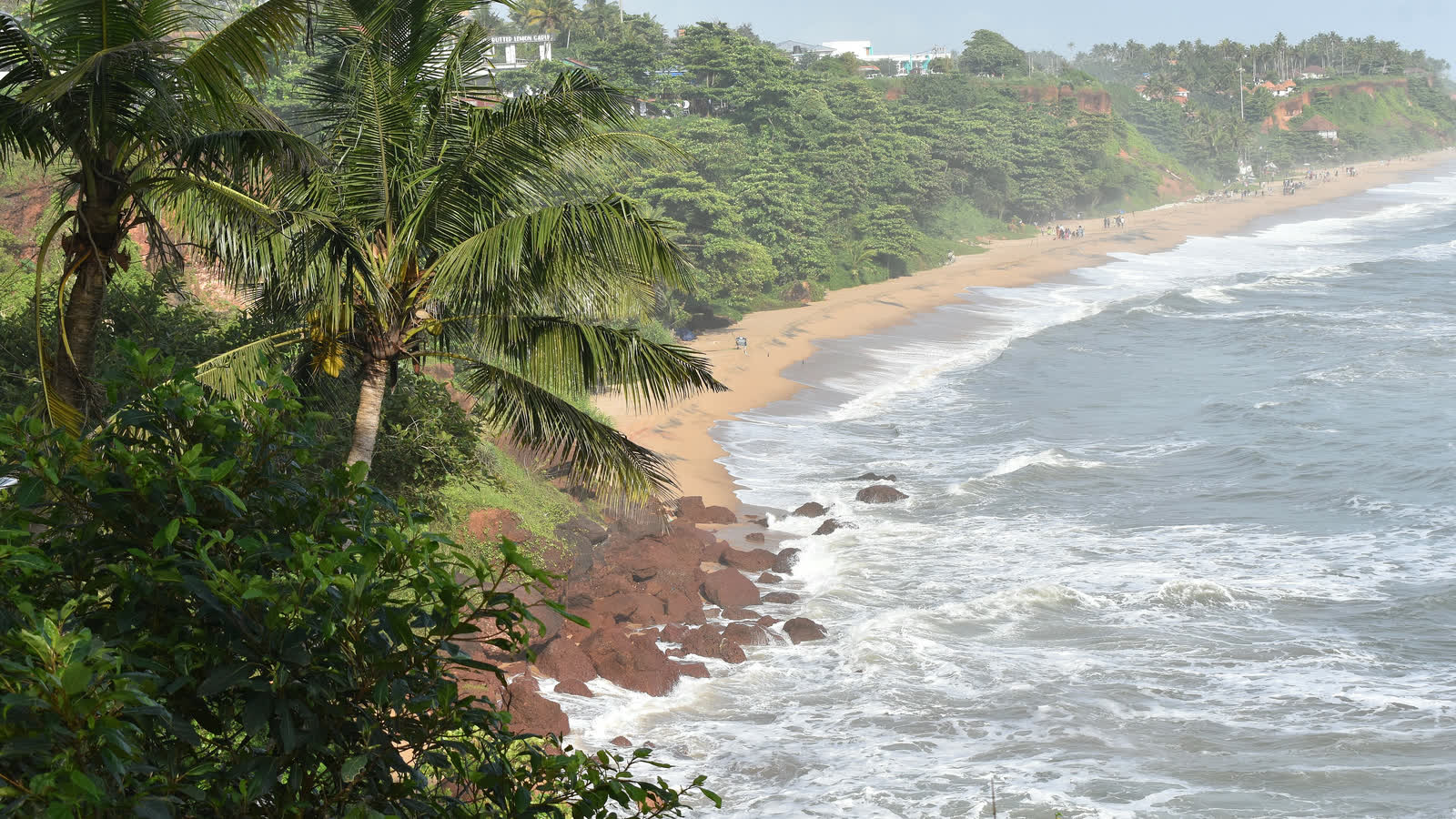 Palm trees on the red cliff at Varkala, looking down on a long sandy beach and white surf, with more cliffs and trees along the coast