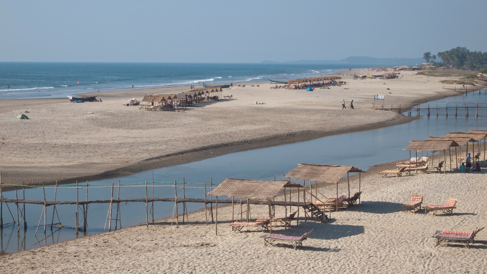 Mandrem beach in North Goa, with beach huts and sun loungers beside a river mouth and a bamboo footbridge