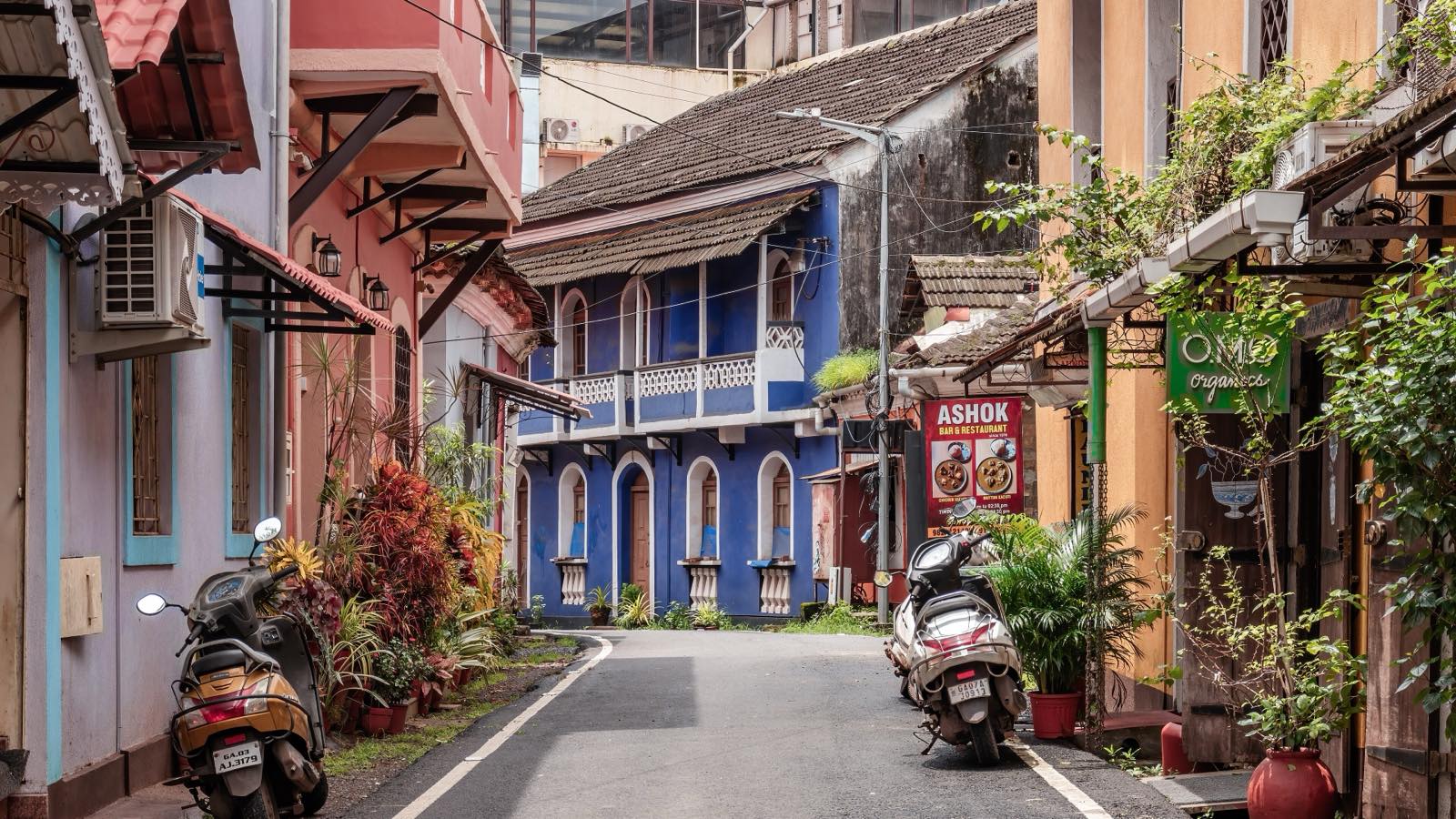 A quiet lane in Fontainhas with a blue two-storey Portuguese-era house at the end, pink and ochre houses on either side and scooters parked by the kerb