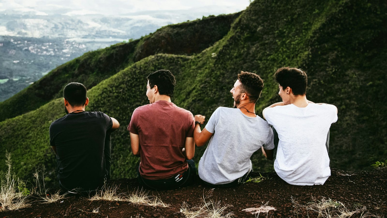 Four friends sitting side by side on a grassy ridge, laughing, with green hills and a valley below