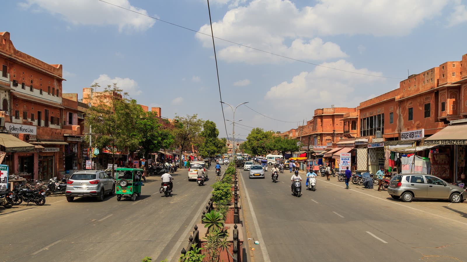 A wide old-city street lined with terracotta-pink buildings, shopfronts and awnings, with scooters and cars in the road