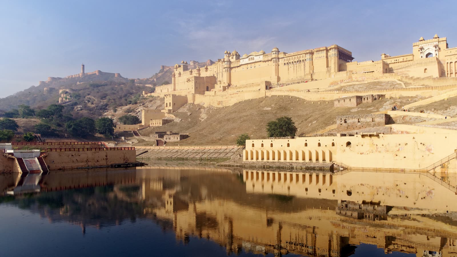 Amer Fort's sandstone walls on the hillside above Maota Lake, reflected in the still water, with Jaigarh Fort on the ridge behind