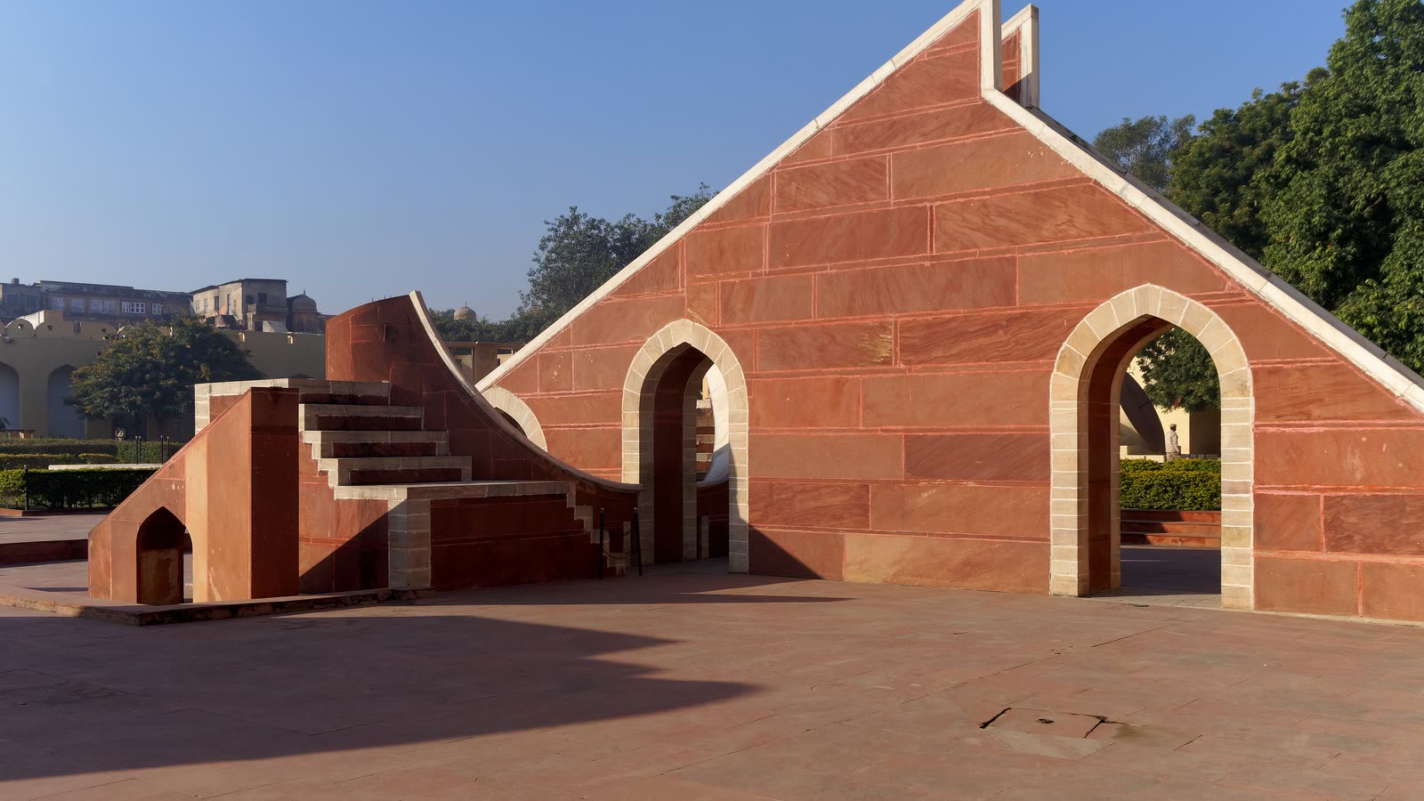 Red sandstone instruments at Jantar Mantar in morning light: a steep triangular wall with arched openings and a curved staircase beside it