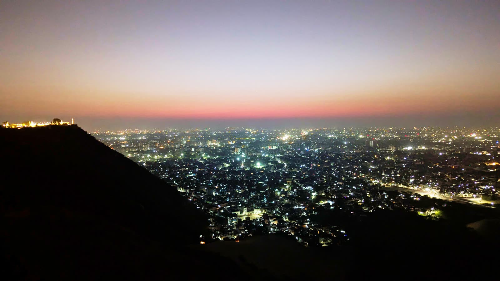 The lights of Jaipur spread across the plain below a dark ridge just after sunset, with a lit-up fort on the ridgetop at the left