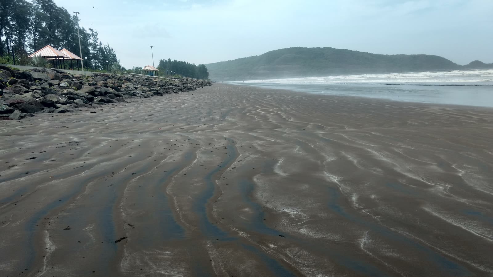 Ripples of wet dark sand on Shrivardhan beach at low tide, with a green hill behind