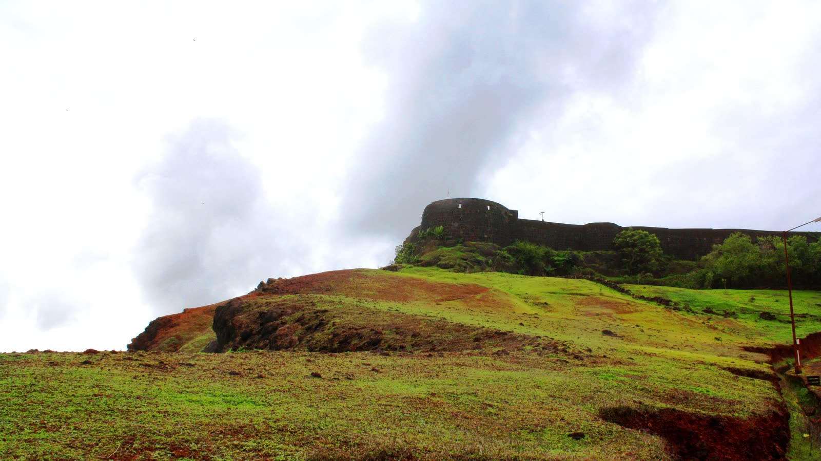 The black stone walls of Ratnadurg Fort on a green hill above red earth, under monsoon clouds