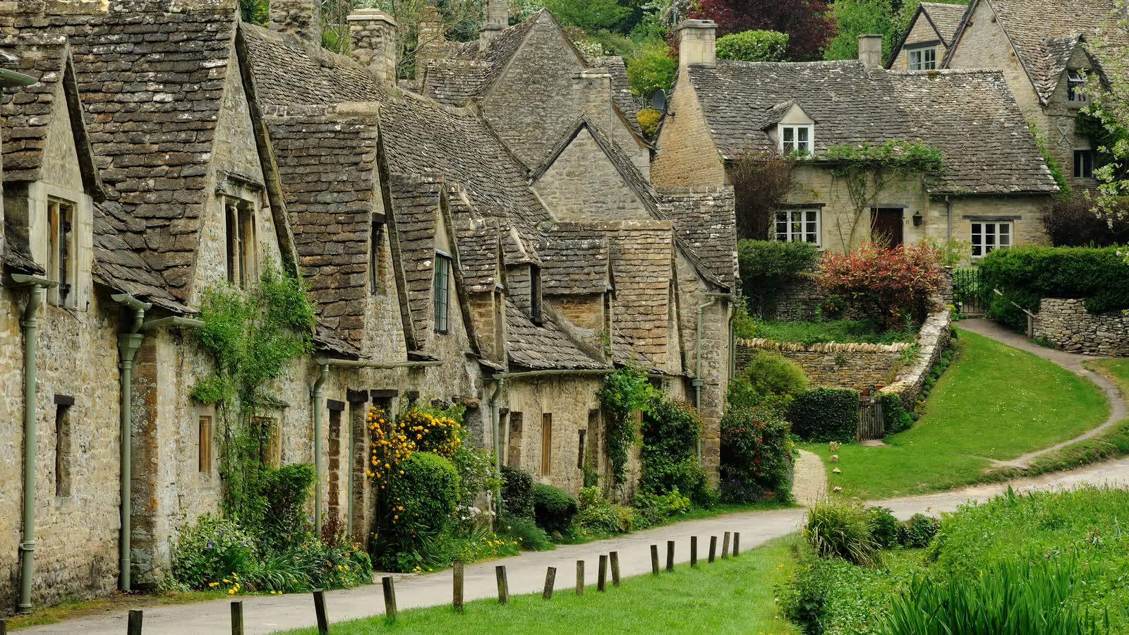 A row of old stone cottages with steep roofs along a path and green bank in Bibury
