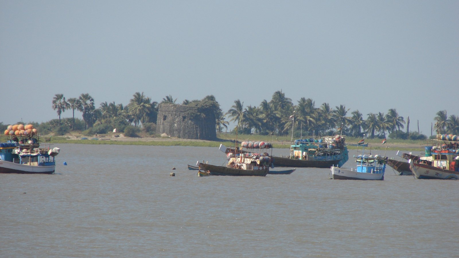 Fishing boats moored in front of Arnala island, with the fort's stone bastion among palm trees