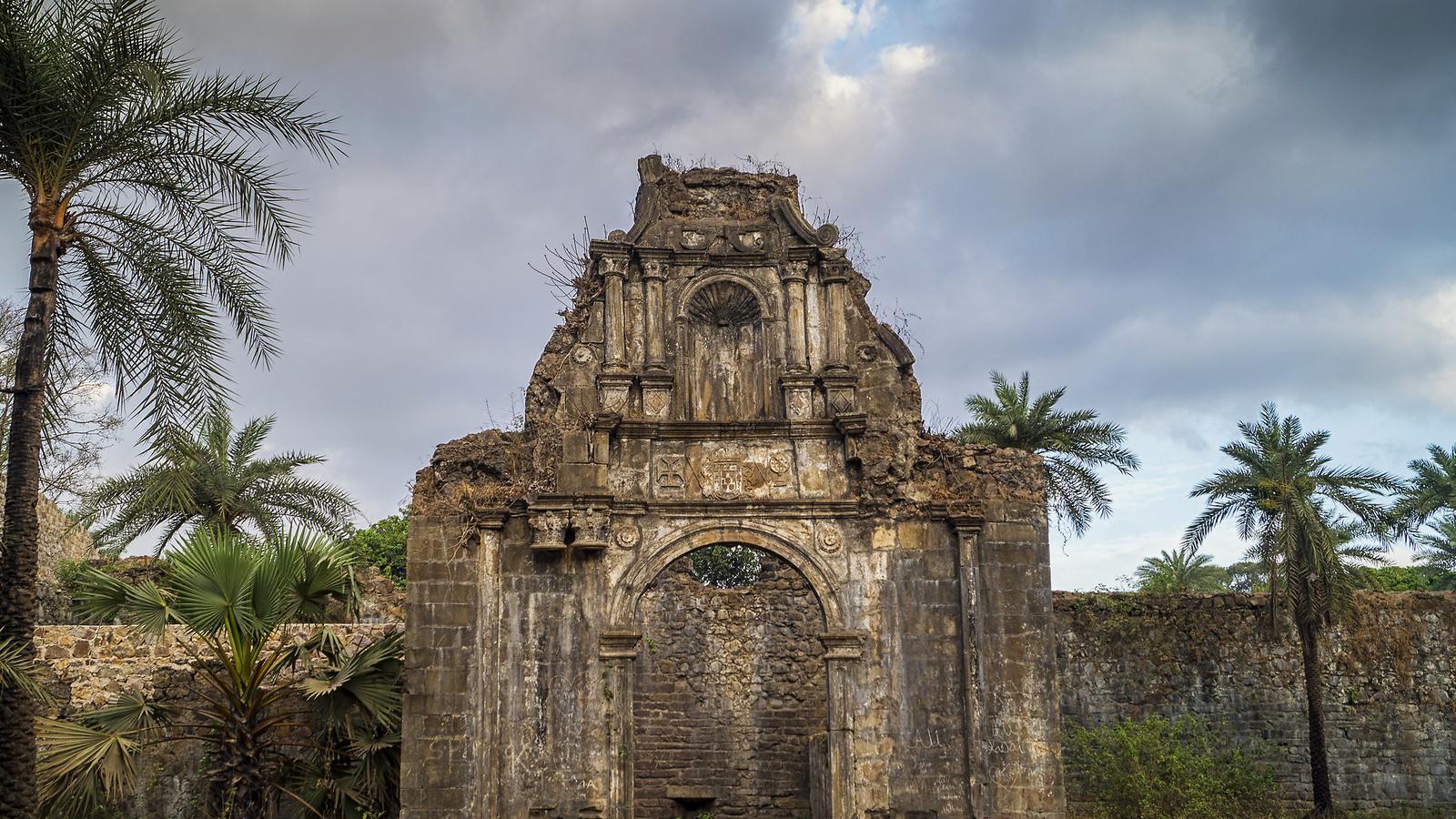 The carved stone gateway of the citadel at Vasai Fort, weathered and overgrown, framed by palm trees