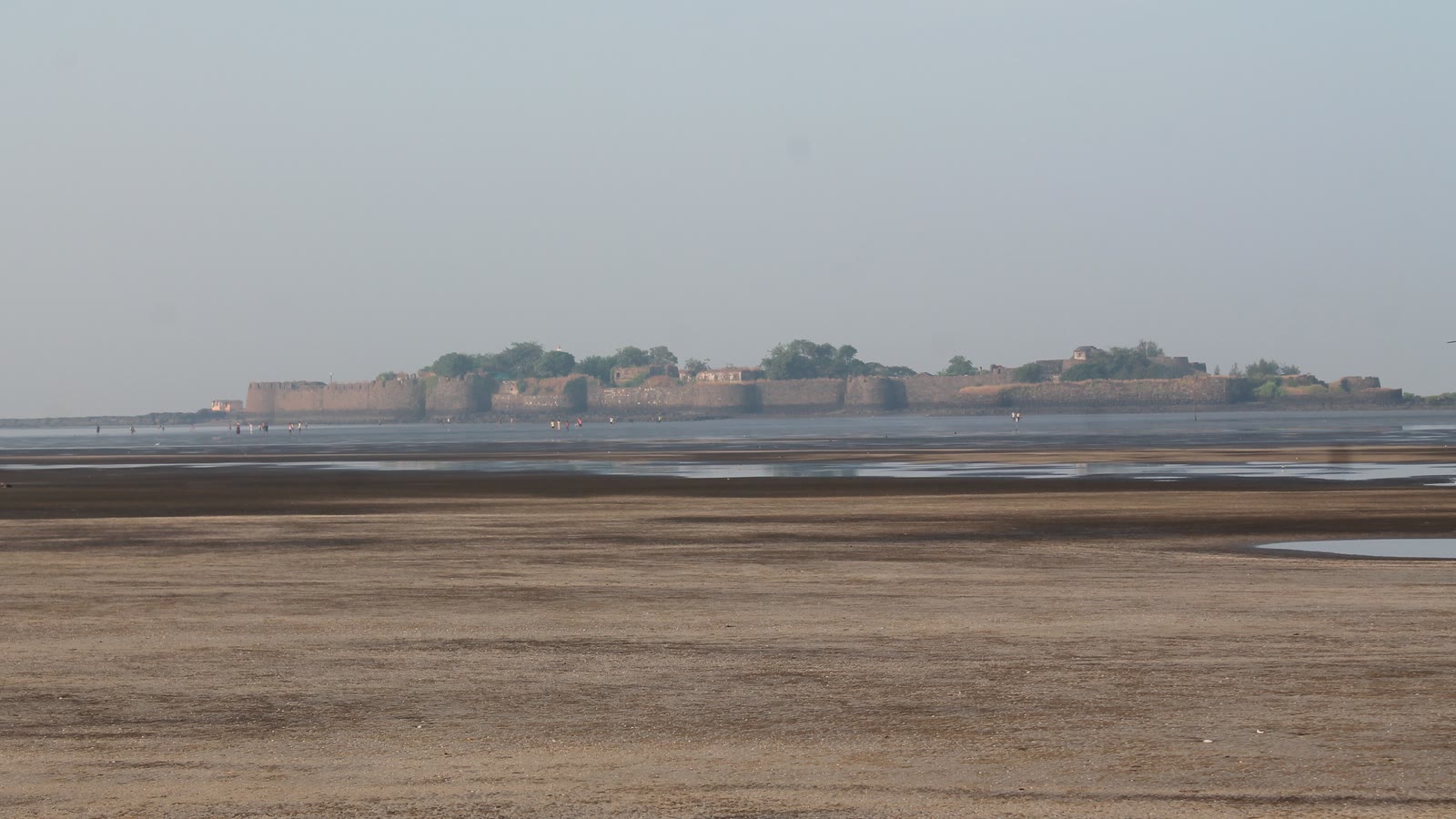 The long stone walls of Kolaba Fort across a wide stretch of wet sand at low tide, with people walking out towards it