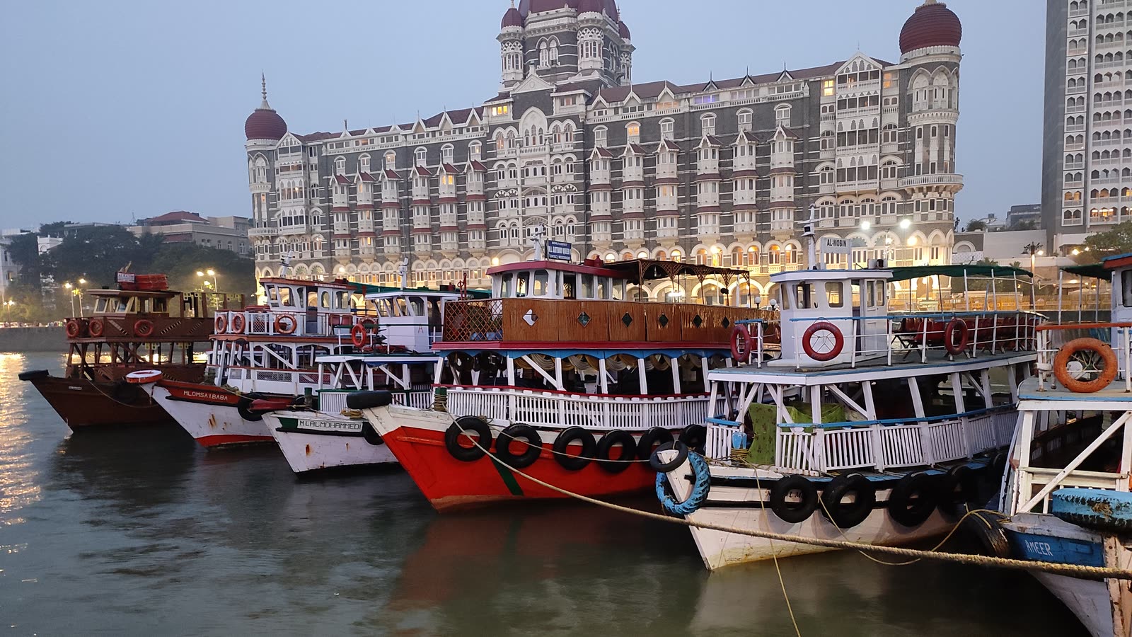 Wooden ferry boats moored by the Gateway of India at dusk, with the Taj Mahal Palace hotel behind