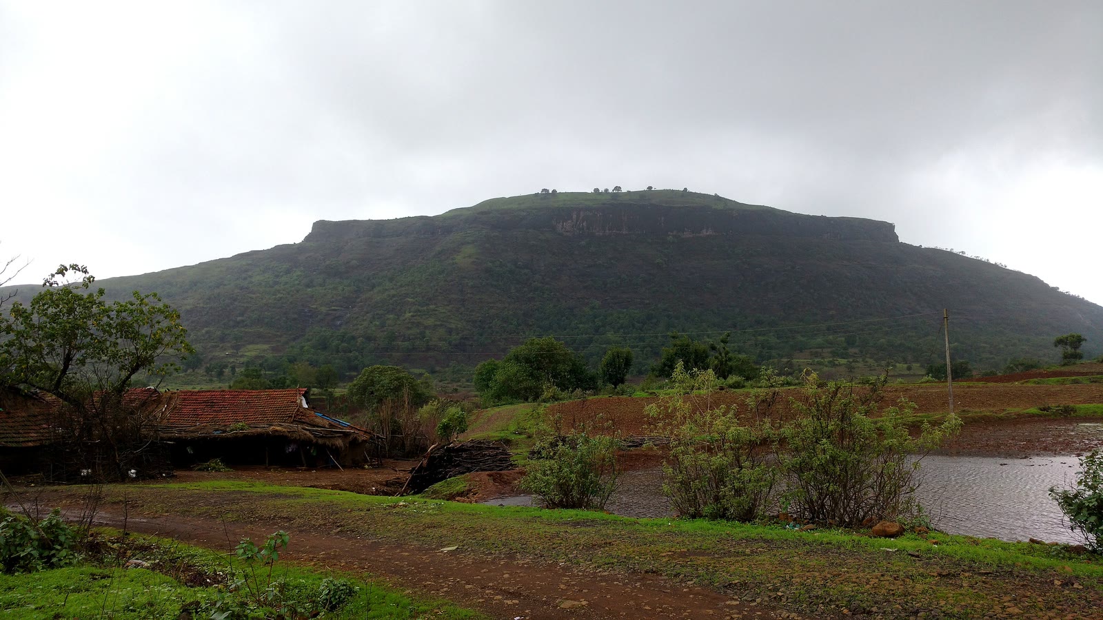 The flat-topped hill of Tringalwadi Fort under monsoon clouds, with a tiled farmhouse and a pond in front
