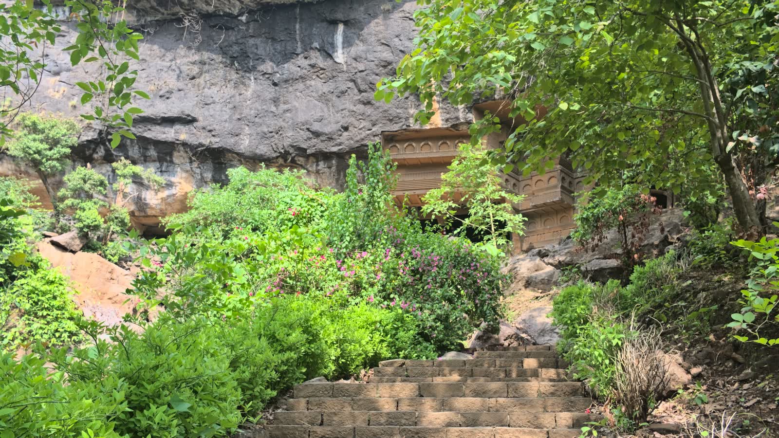 Stone steps leading up through green shrubs to the carved front of the Kondana caves under a rock face