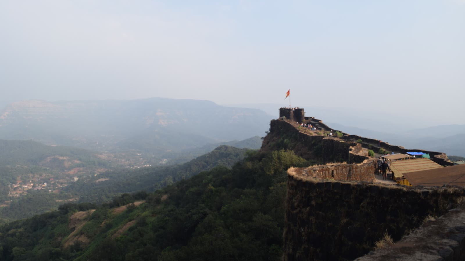 The stone ramparts of Pratapgad Fort on a forested ridge, with a saffron flag and hazy valleys beyond