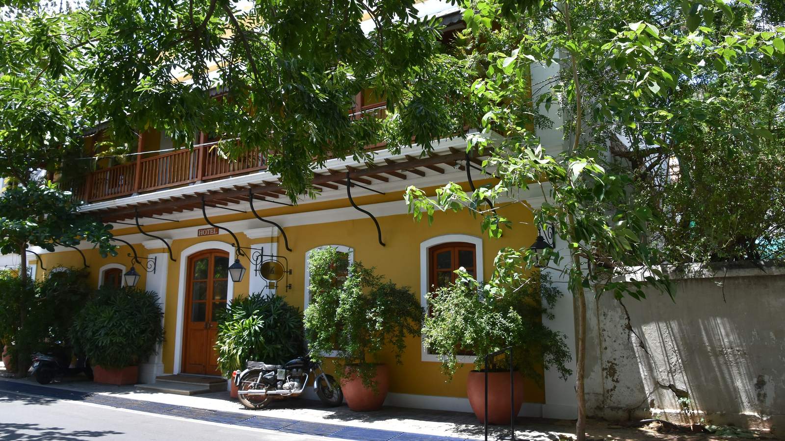 A yellow colonial house with wooden balconies, arched windows and potted plants, under shady trees in Pondicherry's French Quarter