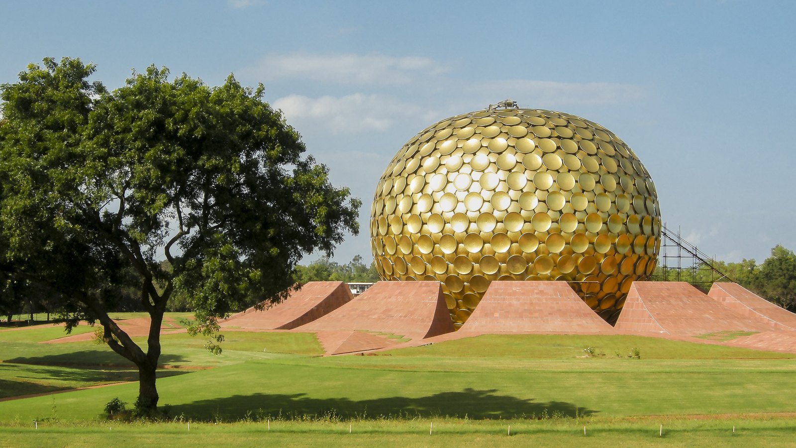 The Matrimandir, a large sphere covered in golden discs, rising from red stone mounds on a green lawn, with a tree in the foreground