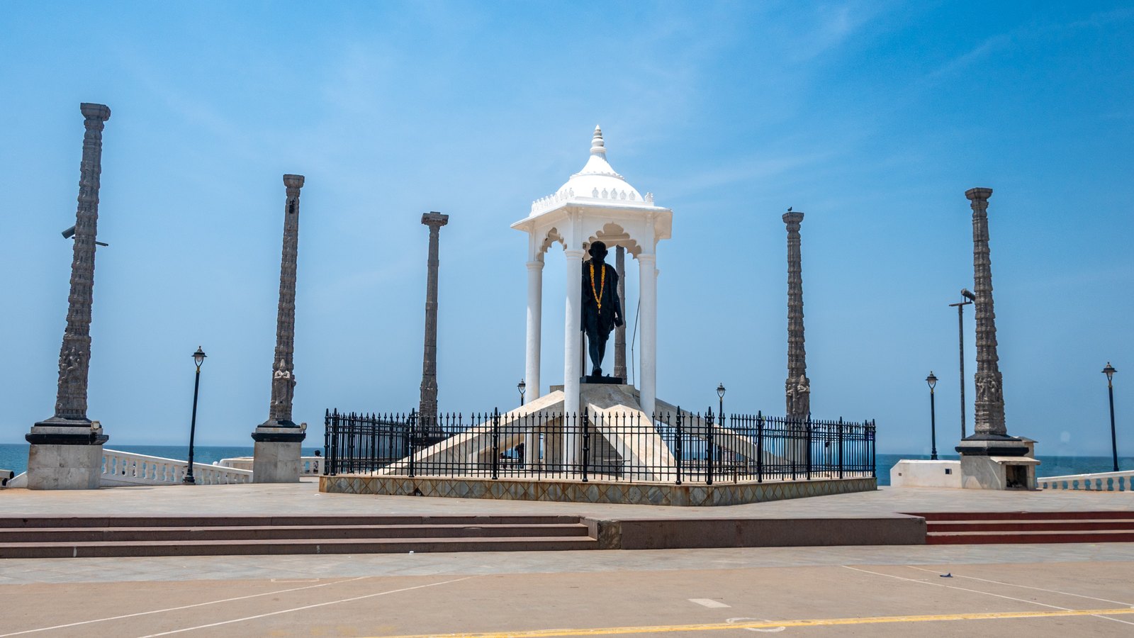 The statue of Mahatma Gandhi under a white canopy on the Promenade, ringed by tall carved stone pillars, with the sea behind