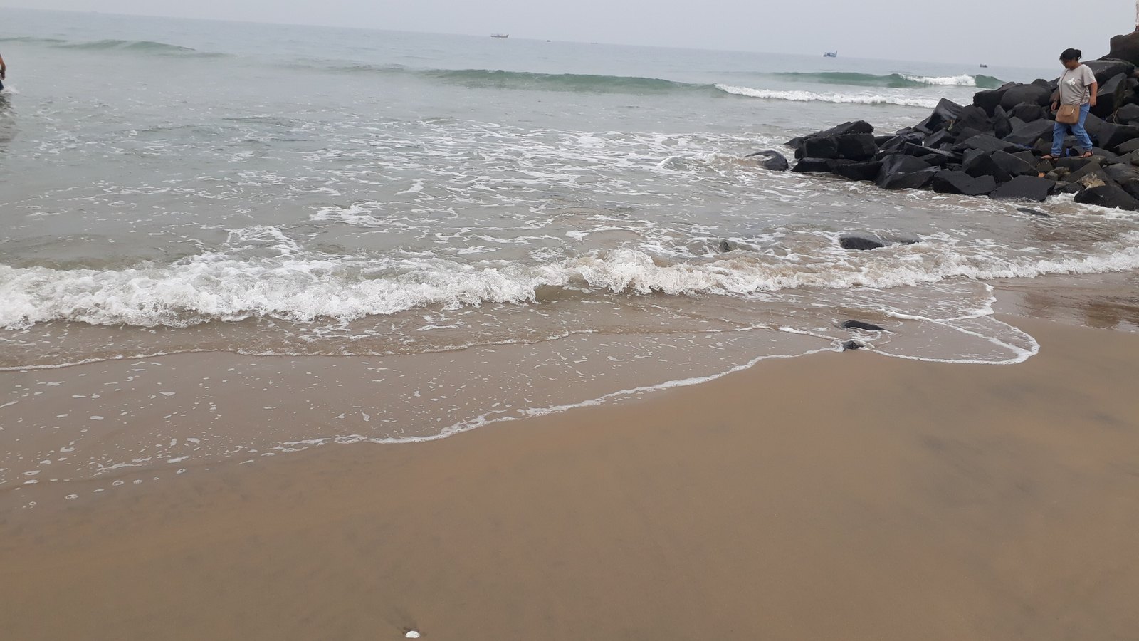 Waves running up a wide sandy beach under a grey sky, with a line of black boulders on the right