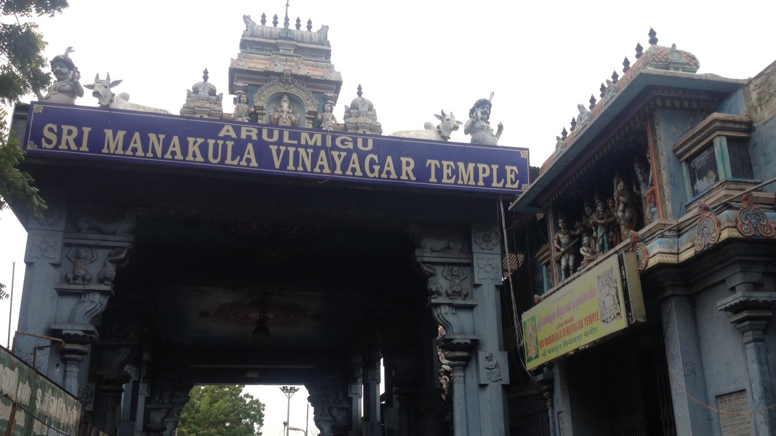 The south entrance of Manakula Vinayagar Temple, a carved stone gateway under a blue sign with the temple's name and painted figures on the roof
