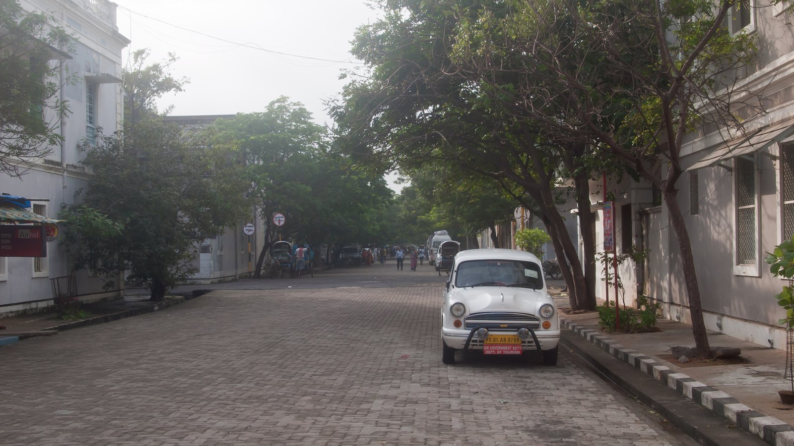 A quiet, tree-lined White Town street paved with grey blocks, with pale colonial buildings on both sides and a white Ambassador car parked at the kerb