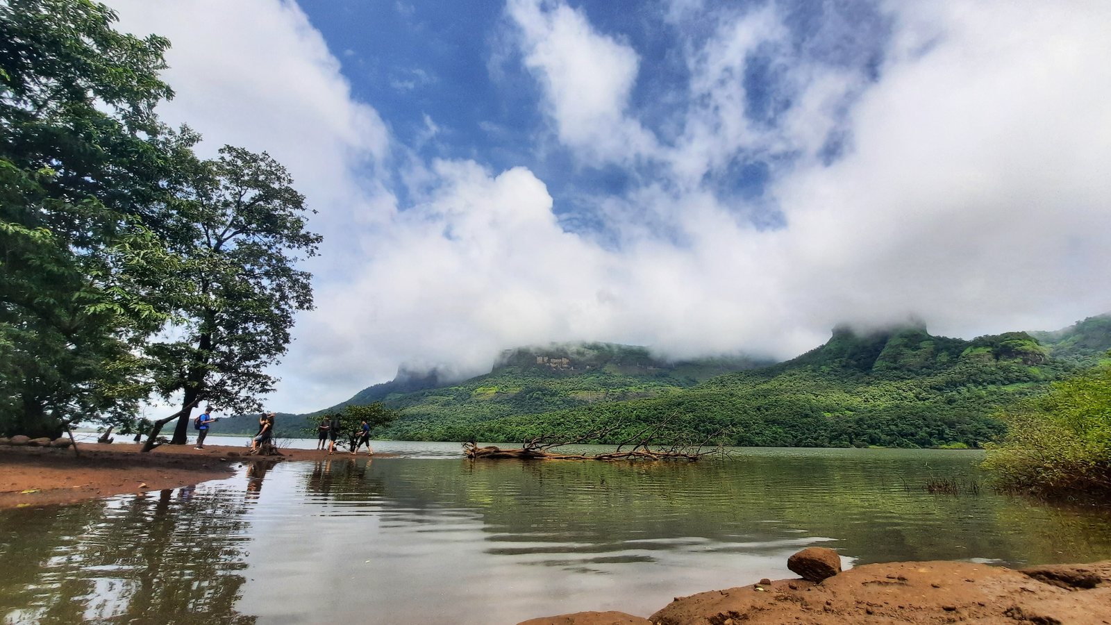 The calm Kundalika river with a sandy bank, trees and a forested hill under white monsoon clouds