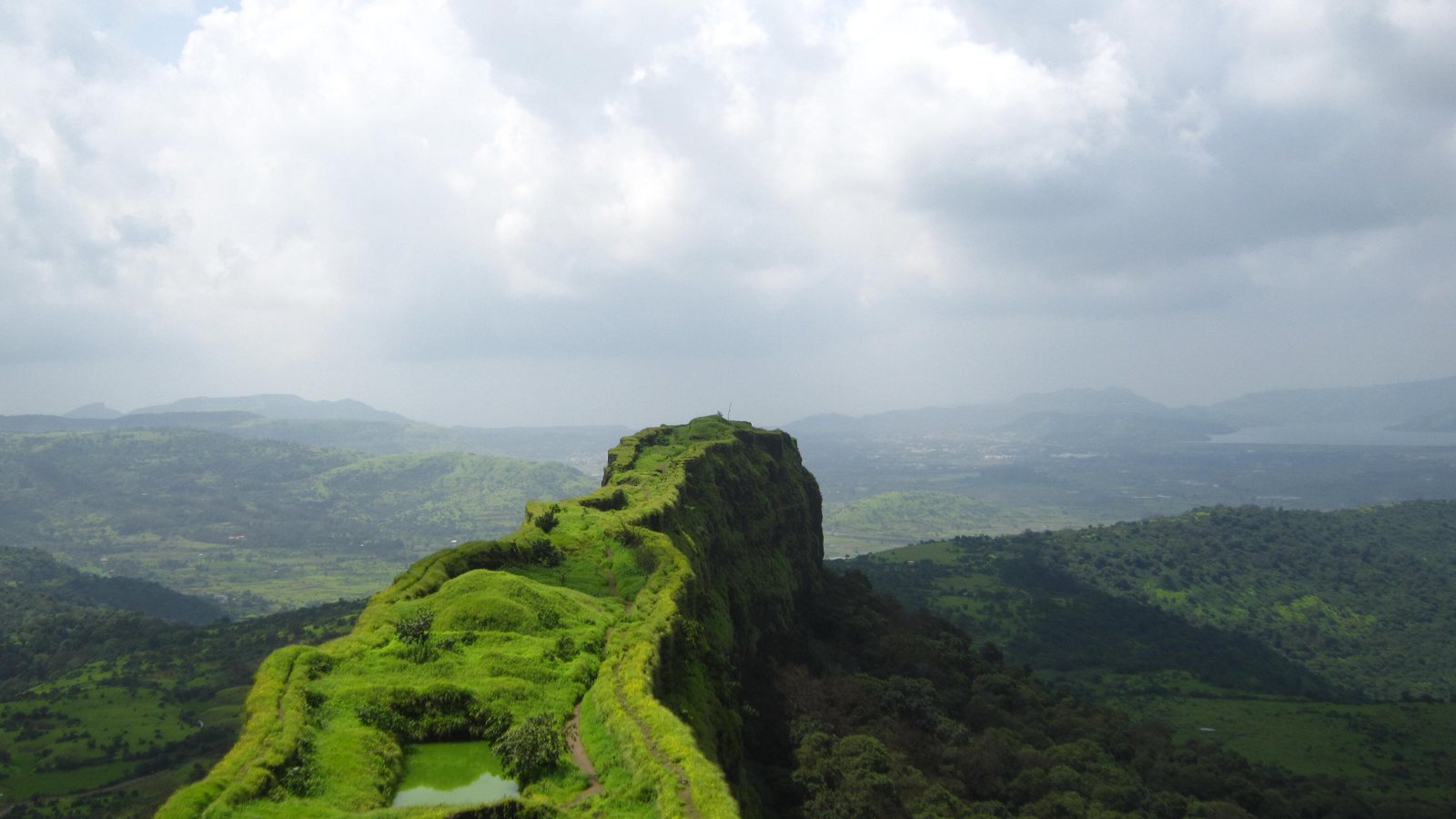 The narrow, grassy spur of Lohagad Fort running out over green valleys under monsoon clouds
