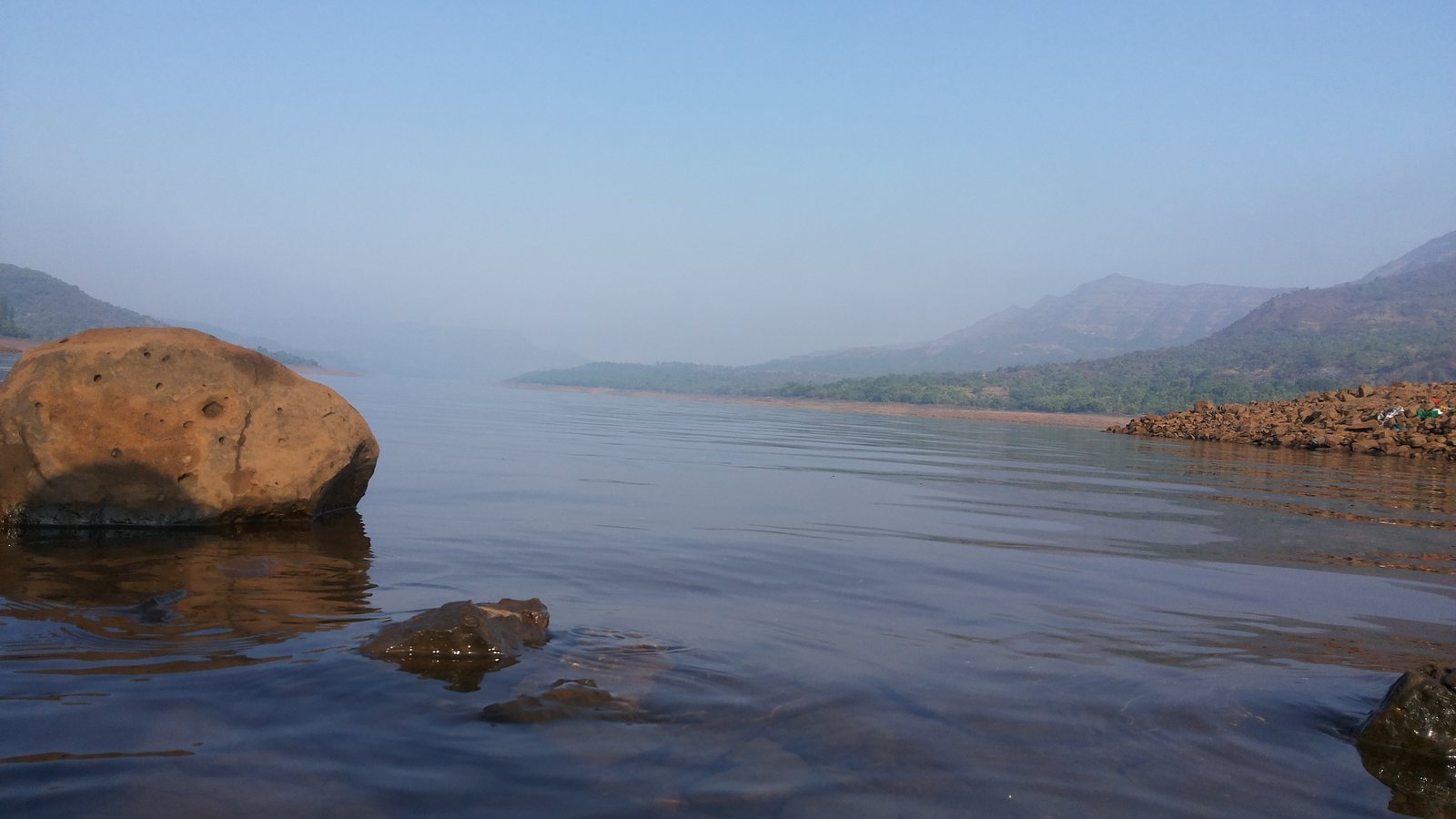 Still water on Mulshi lake with a large boulder in the foreground and hazy hills behind