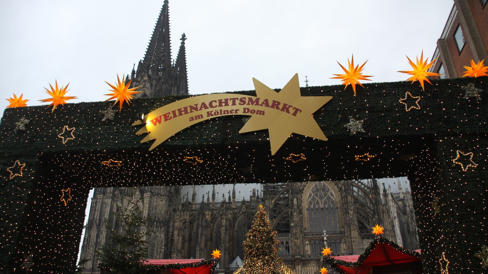A green entrance arch with gold stars and a sign reading Weihnachtsmarkt am Kölner Dom, with the cathedral's spires rising behind it