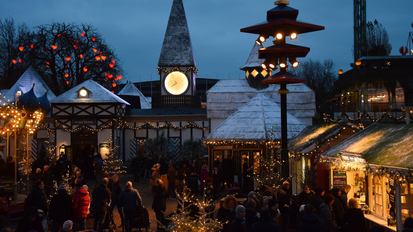 Christmas market huts with snowy roofs and strings of lights around a clock tower in Tivoli Gardens at dusk, with red heart lights in the trees