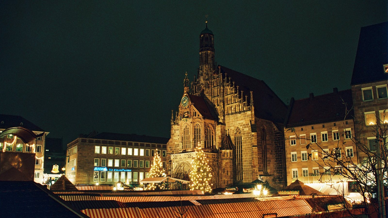 The lit Gothic front of the Frauenkirche at night, with two Christmas trees and the striped roofs of market stalls below