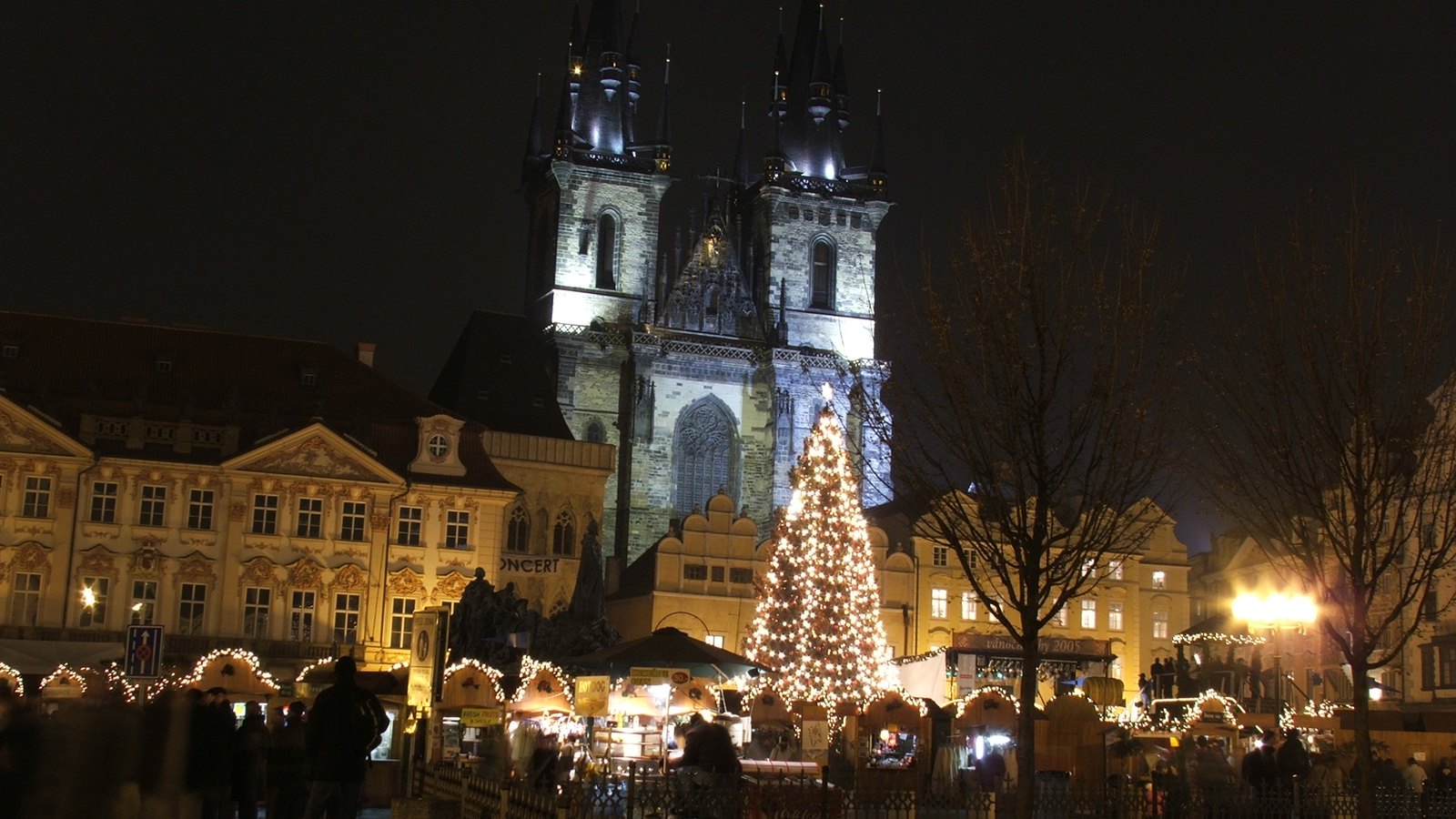The twin spires of Týn Church lit up at night above a Christmas tree and market stalls on Old Town Square in Prague