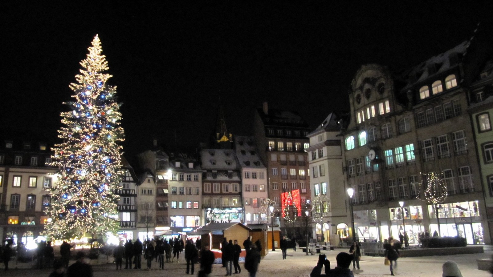 A tall Christmas tree covered in lights in a snowy square in Strasbourg at night, with old townhouses and market chalets behind